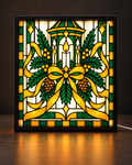 Illustrative framed preview of a golden Christmas lantern candle surrounded by green evergreen sprigs, pinecones and a sweeping golden bow inside a slim black illuminated tabletop frame
