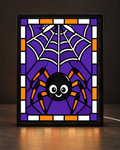 Happy round black spider with orange legs hangs below white web against purple stained-glass mosaic in matte-black illuminated frame; illustrative digital framed preview